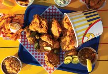 Fried chicken spilling out of a bucket, surrounded by iced team, pickles, crinkle fries, and more