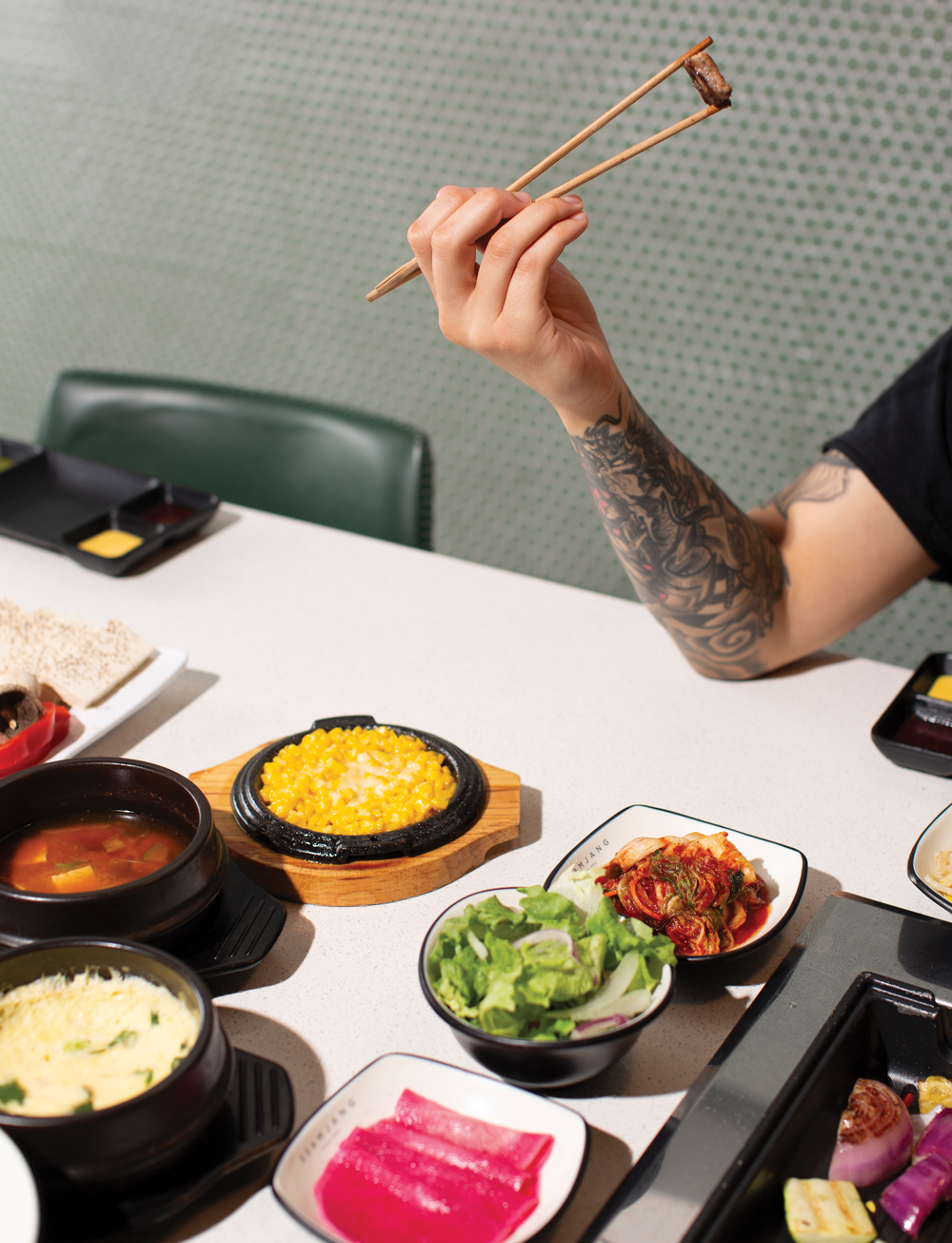 A person with a tattooed arm holding a piece of meat between chop sticks with a spread of dishes in front of them at SsamJang Korean BBQ