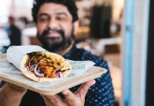 A man smiling in the background while he holds a hot buttered naan wrap