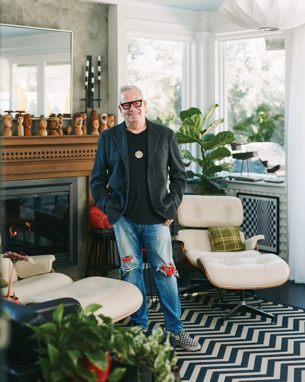 Grant Henry stands in his eclectic home amidst large indoor plants, mid century, white and wooden chairs, and small wooden busts above the fireplace. The black and white chevron rug compliments the checkered wall artwork behind him.
