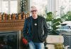 Grant Henry stands in his eclectic home amidst large indoor plants, mid century, white and wooden chairs, and small wooden busts above the fireplace. The black and white chevron rug compliments the checkered wall artwork behind him.