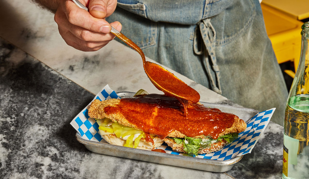 A man using a spoon to pour chili oil on top of a grilled fish sandwich