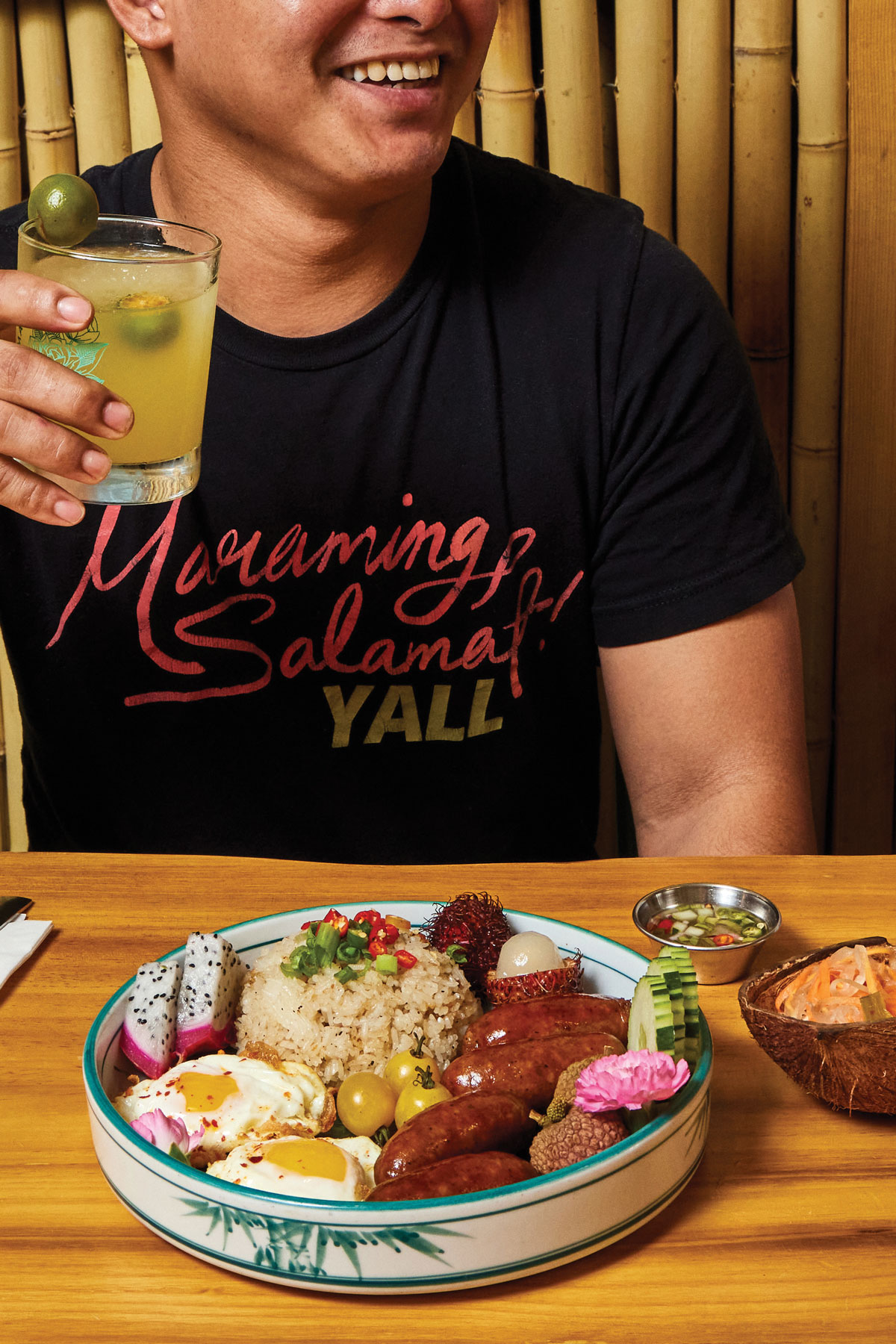 A man smiles while holding a cocktail, a longsilog bowl sits in front of him