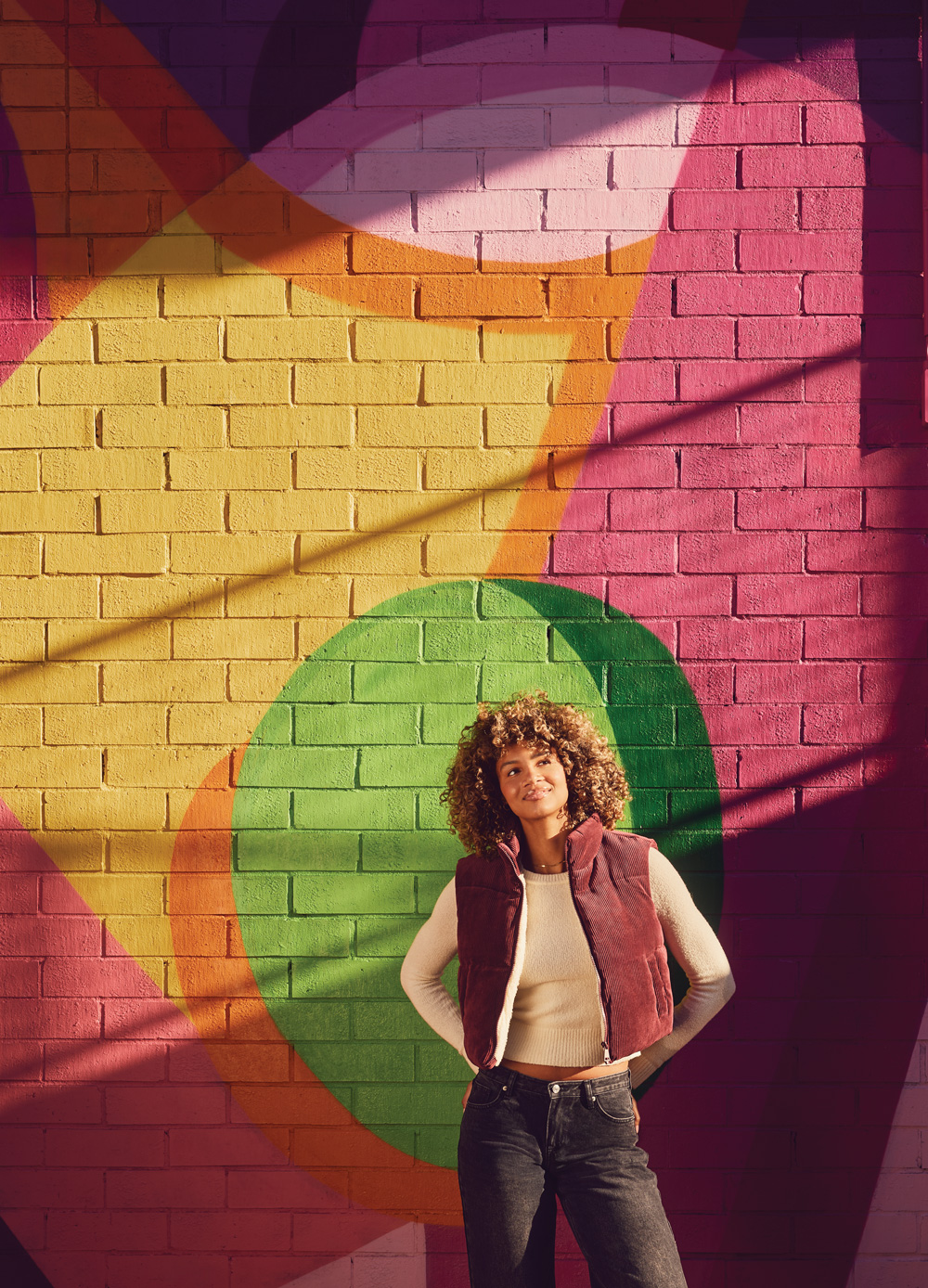 a model smiles in front of a pink, yellow, orange, and green mural