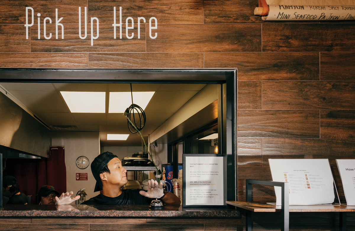a man waits at a takeout window with the words "Pick Up Here" written above on the wall