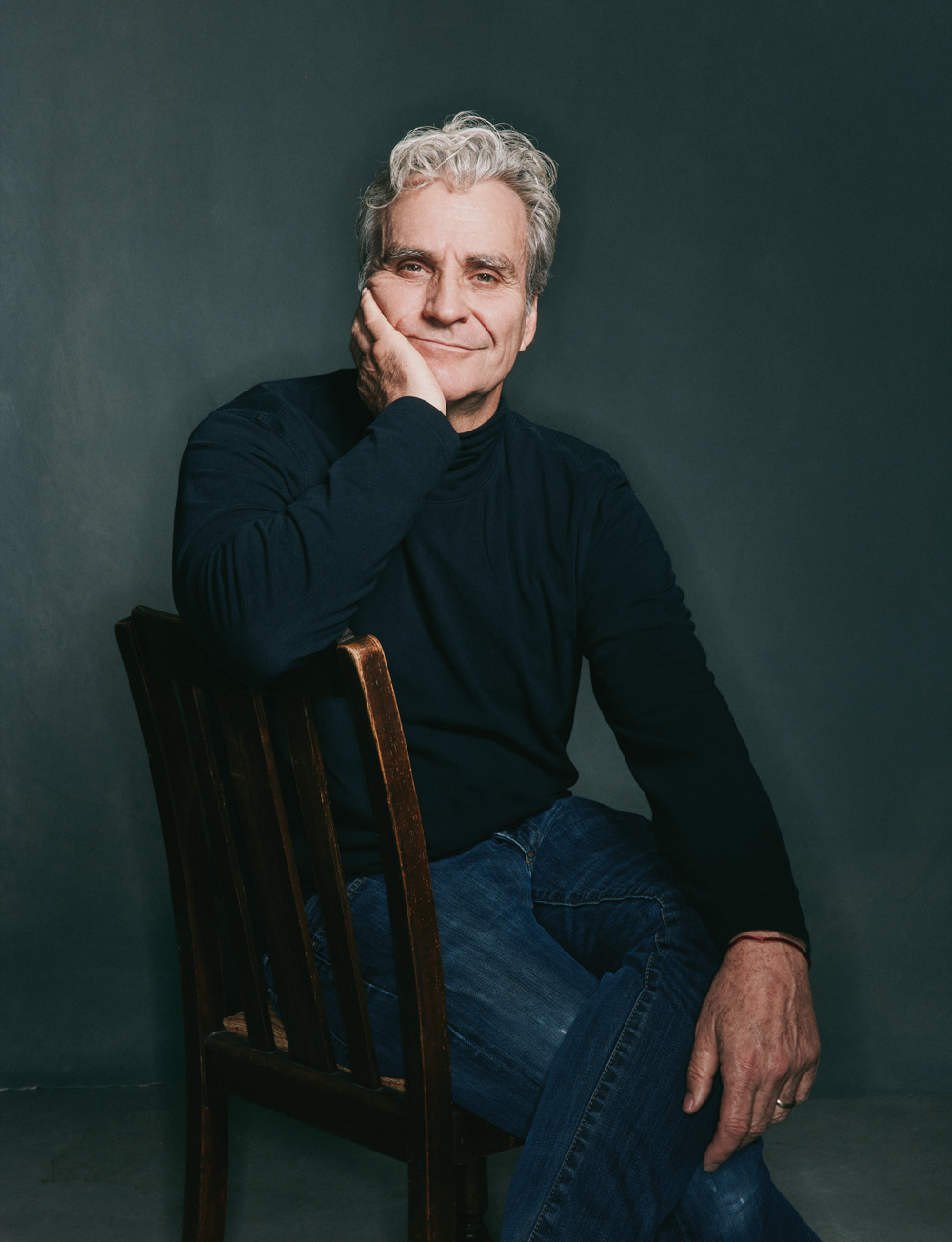 Tom Junod sits in a chair against a dark grey/blue backdrop, wearing a dark turtleneck and jeans