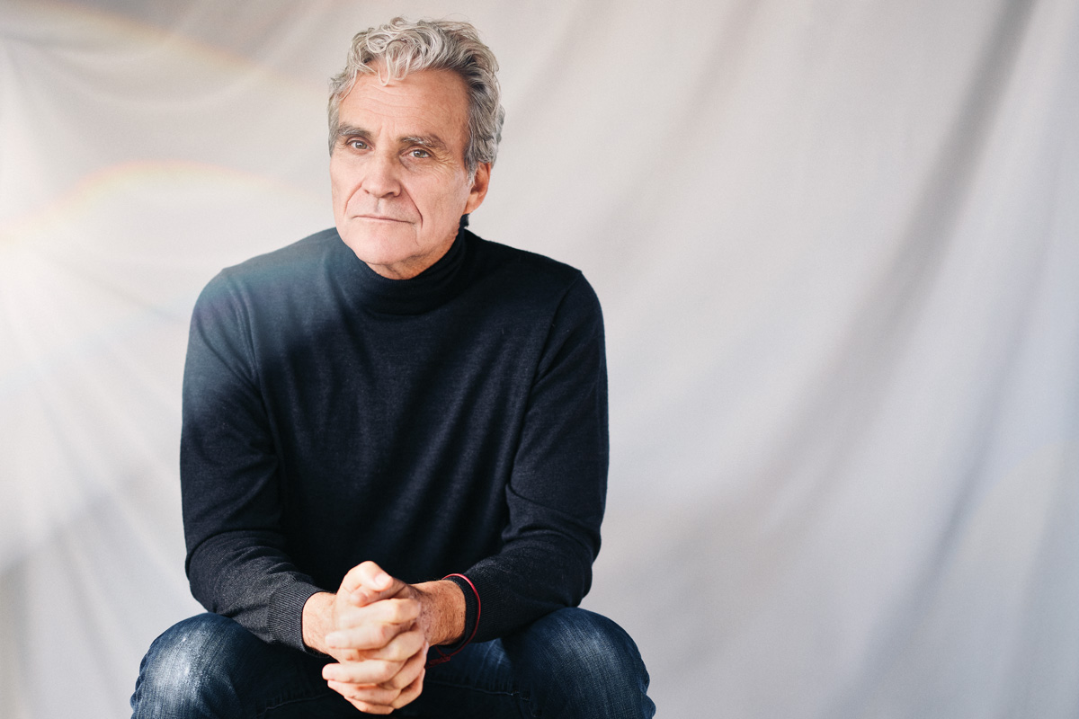 Tom Junod sits in a chair against a white backdrop, wearing a dark turtleneck and jeans