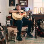 Kevn Kinney holds a guitar in his home, along with his three dogs surrounding him
