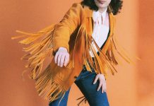 a person models blue jeans, a white button up, and a brown fringe suede jacket against a burnt orange backdrop