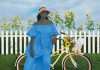 a woman in a blue dress stands by her bike with a white dog in the basket and a white picket fence in the background